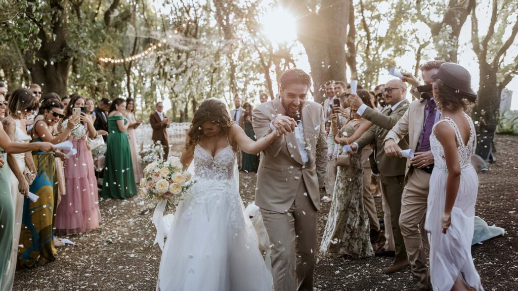 Simone, con il suo abito color sabbia, e Chiara vengono accolti da petali lanciati dagli invitati in un’atmosfera di gioia.