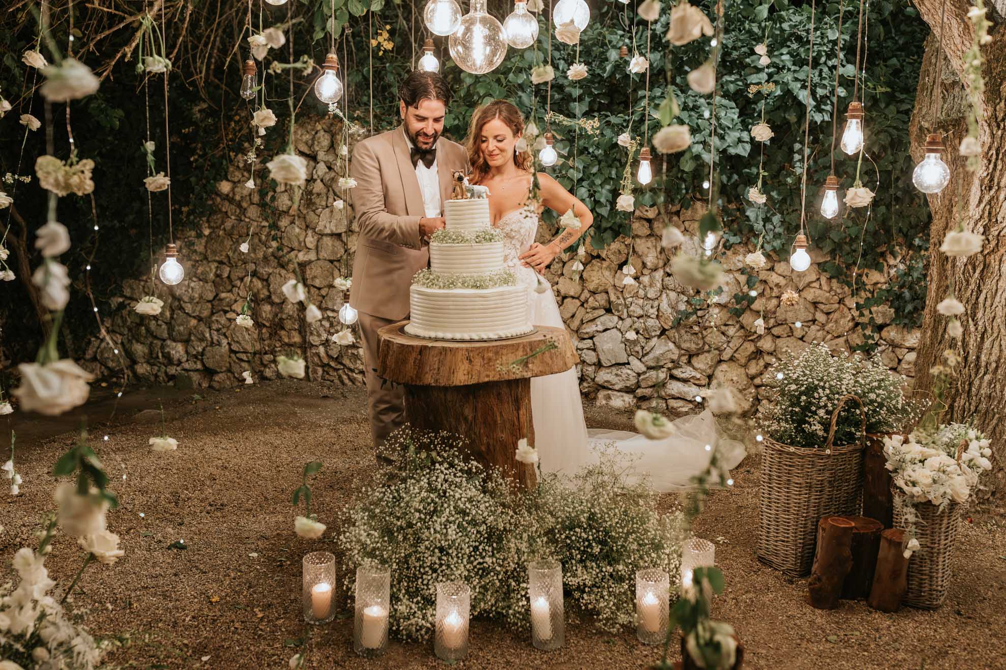 Simone, in un elegante abito color sabbia, taglia la torta nuziale accanto a Chiara, in un’atmosfera romantica tra luci e fiori.