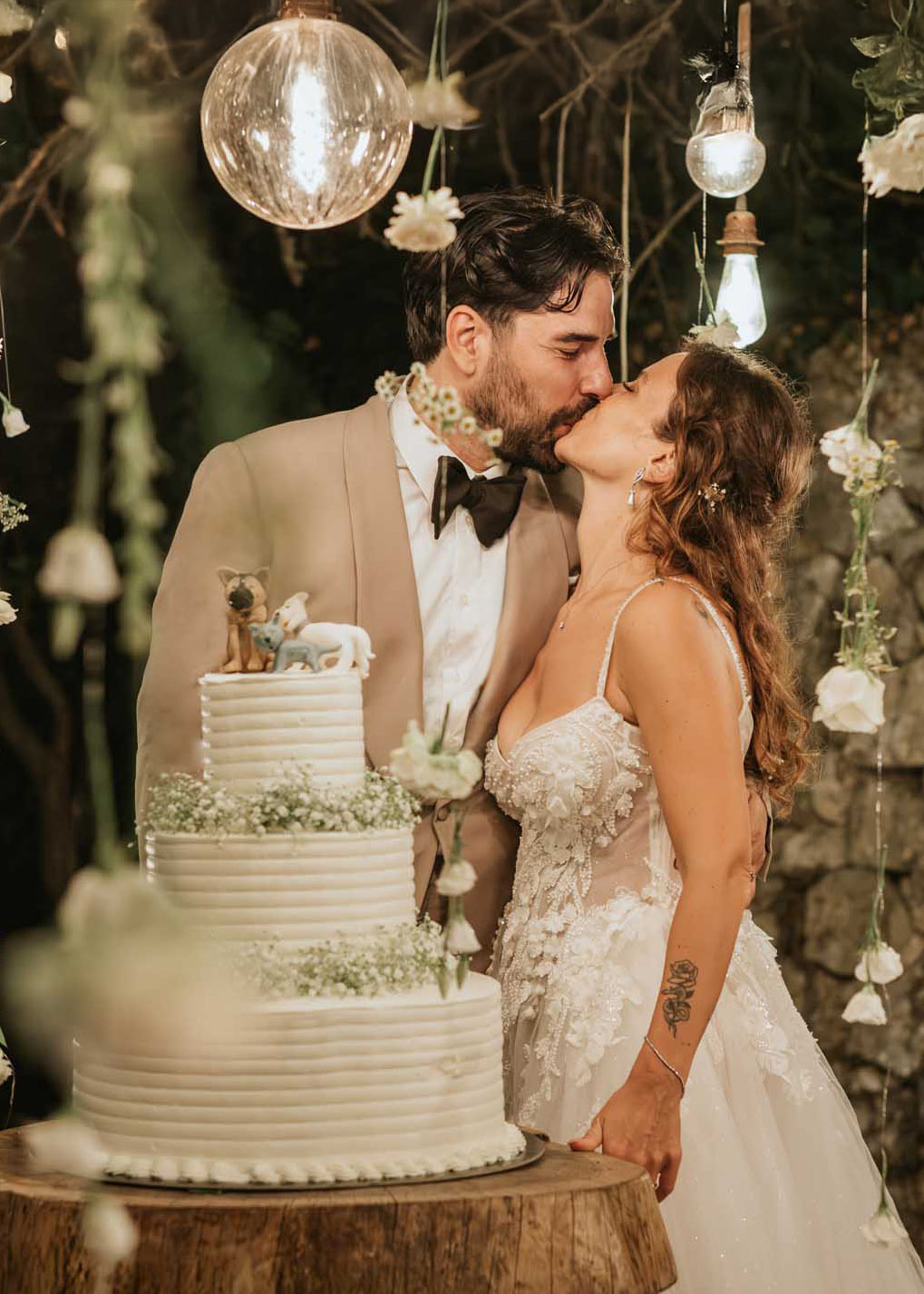 Simone, con un abito sartoriale sabbia, e Chiara si baciano accanto alla torta nuziale decorata con dettagli floreali.